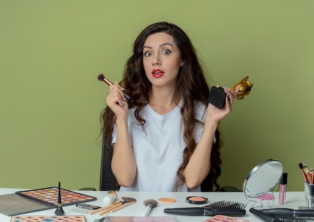 Impresionada niña bonita sentada en la mesa de maquillaje con herramientas de maquillaje con pincel en polvo y copa ganador aislado sobre fondo verde oliva