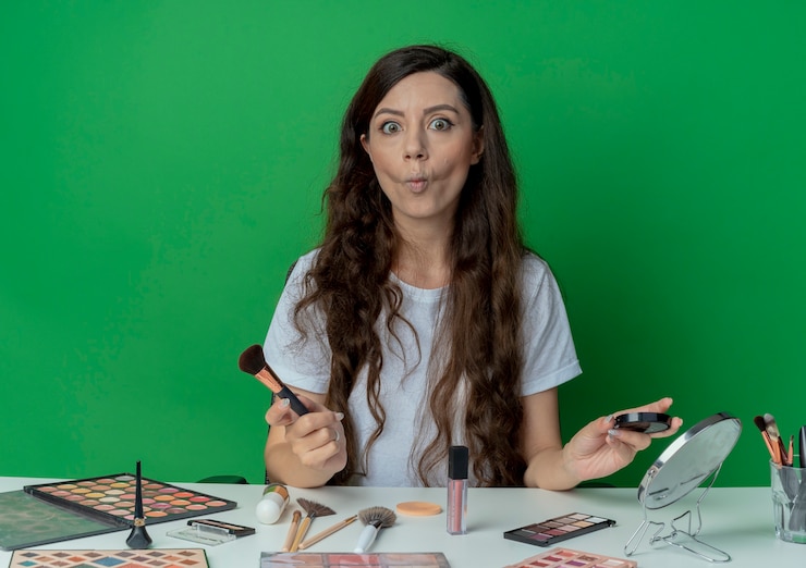Foto gratuito impresionada niña bonita sentada en la mesa de maquillaje con herramientas de maquillaje con pincel de rubor y rubor aislado sobre fondo verde