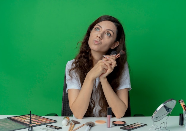 Foto gratuita impresionada niña bonita sentada en la mesa de maquillaje con herramientas de maquillaje con pinceles en polvo y rubor mirando hacia arriba aislado sobre fondo verde