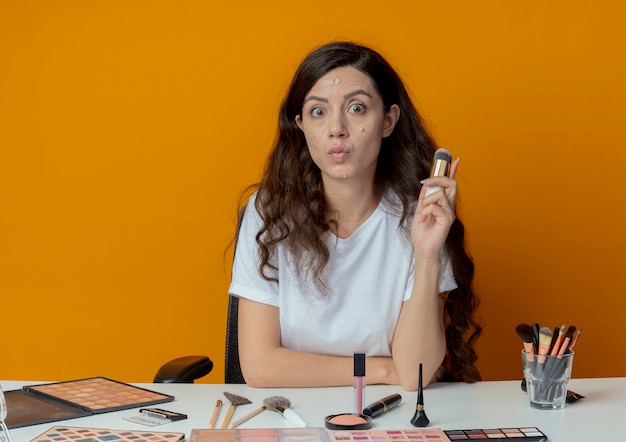 Impresionada niña bonita sentada en la mesa de maquillaje con herramientas de maquillaje sosteniendo un cepillo de base con crema de base puesta en su rostro aislado sobre fondo naranja