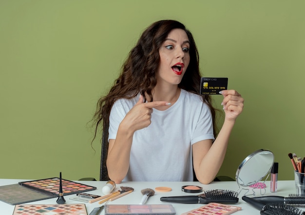 Impresionada niña bonita sentada en la mesa de maquillaje con herramientas de maquillaje sosteniendo mirando y apuntando a la tarjeta de crédito en el espacio verde oliva