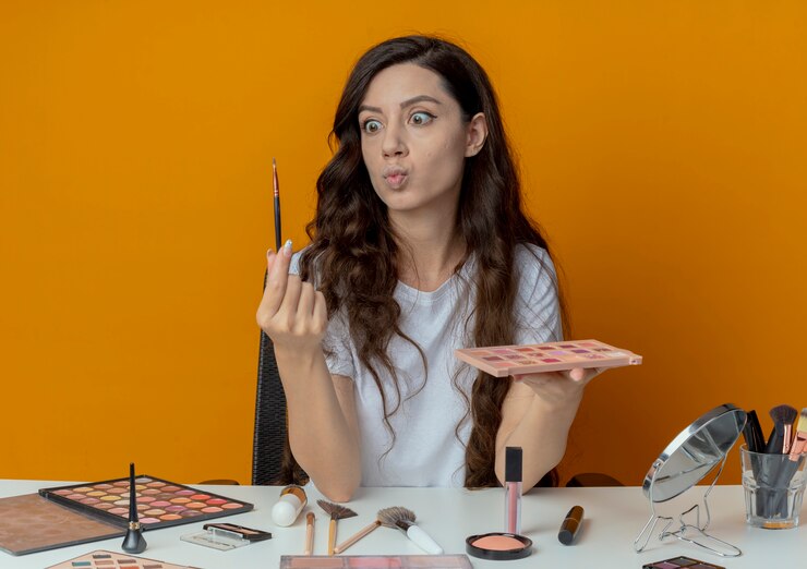 Foto gratuito impresionada niña bonita sentada en la mesa de maquillaje con herramientas de maquillaje sosteniendo la paleta y el pincel de sombra de ojos y mirando el pincel aislado sobre fondo naranja