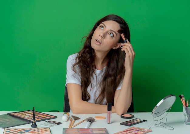 Impresionada niña bonita sentada en la mesa de maquillaje con herramientas de maquillaje sosteniendo un pincel de rubor y tocando la frente con él y mirando hacia arriba aislado sobre fondo verde