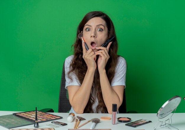 Impresionada niña bonita sentada en la mesa de maquillaje con herramientas de maquillaje sosteniendo pinceles de rubor y polvo y tocando la cara con ellos aislados sobre fondo verde