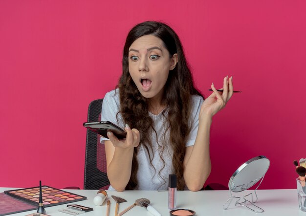 Impresionada niña bonita sentada en la mesa de maquillaje con herramientas de maquillaje sosteniendo teléfono móvil y pincel de maquillaje mirando teléfono aislado sobre fondo carmesí