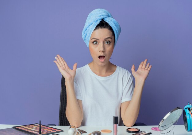 Impresionada niña bonita sentada en la mesa de maquillaje con herramientas de maquillaje y con una toalla de baño en la cabeza mostrando las manos vacías aisladas sobre fondo púrpura