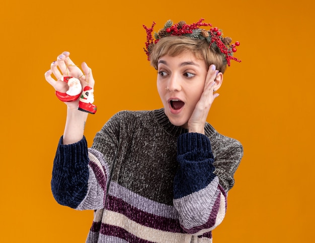 Impresionada niña bonita vistiendo corona de cabeza de navidad sosteniendo y mirando a santa claus adornos de navidad manteniendo la mano en la cara aislada sobre fondo naranja