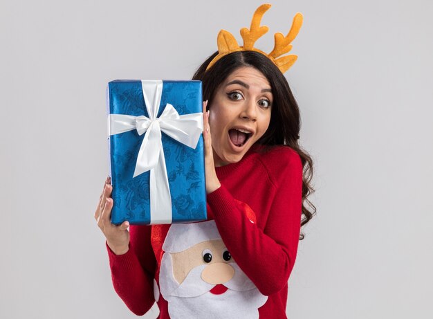 Impresionada niña bonita vistiendo cuernos de reno diadema y suéter de santa claus sosteniendo el paquete de regalo de navidad mirando