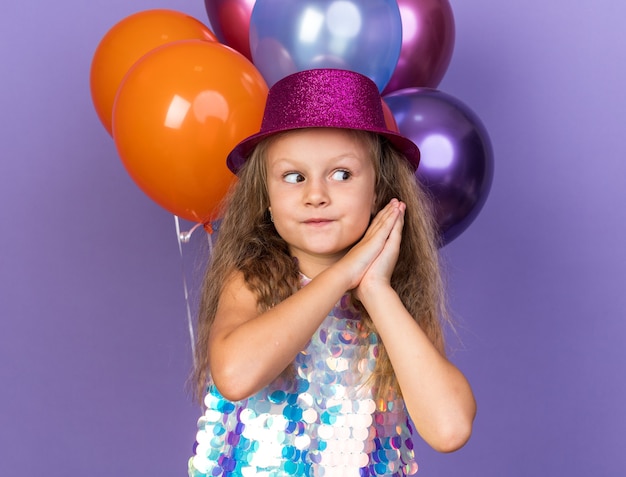 Impresionada niña rubia con sombrero de fiesta violeta tomados de la mano juntos y mirando al lado de pie con globos de helio aislado en la pared púrpura con espacio de copia