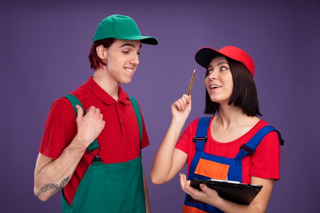 Impresionada pareja joven en uniforme de trabajador de la construcción y gorra chica sosteniendo lápiz y portapapeles mirando chico apuntando hacia arriba con lápiz chico mordiendo el labio mirando portapapeles tocando su uniforme