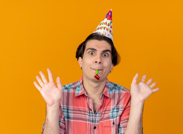 Impresionado apuesto hombre caucásico vestido con gorro de cumpleaños se encuentra con las manos levantadas soplando el silbato de fiesta