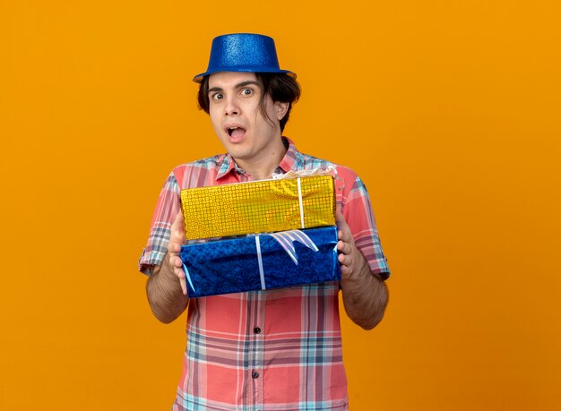 Impresionado apuesto hombre caucásico vestido con gorro de fiesta azul tiene cajas de regalo