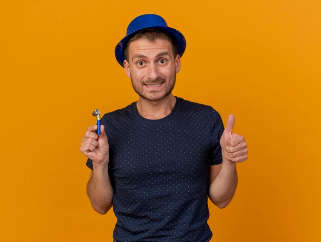 Impresionado apuesto hombre vestido con gorro de fiesta azul Thumbs up y sostiene un silbato de fiesta aislado en la pared naranja con espacio de copia