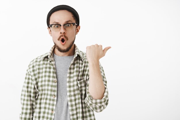 Impresionado y asombrado, un chico guapo y emotivo con barba con gafas y gorro doblando los labios con un sonido increíble, apuntando a la derecha con el pulgar y mirando asombrado.