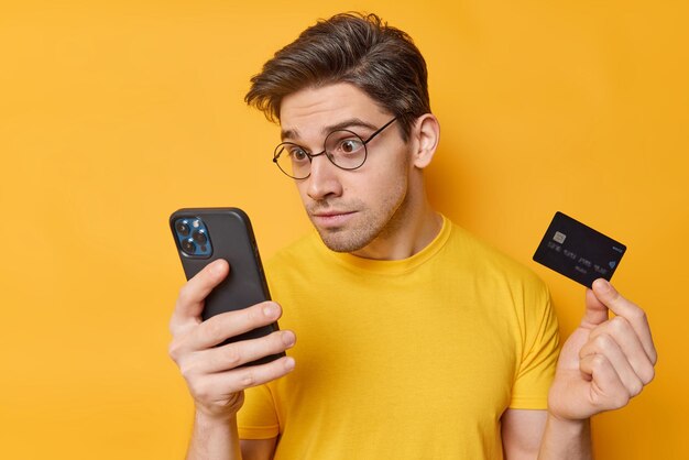 Impresionado, atónito, el hombre brunet mira fijamente la pantalla del teléfono inteligente, sostiene la tarjeta de crédito, reacciona ante descuentos inesperados, usa anteojos redondos, una camiseta informal aislada sobre la aplicación de descargas de fondo amarillo