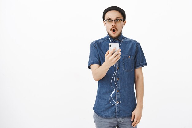 Impresionado chico guapo sin palabras con bigote en anteojos y gorro negro jadeando abriendo la boca con sonido wow mirando después de leer un mensaje impactante en un teléfono inteligente con auriculares