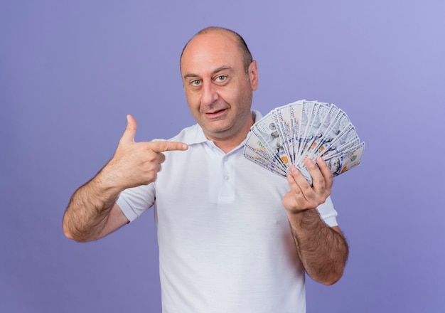 Impresionado empresario maduro casual sosteniendo y apuntando al dinero aislado en púrpura