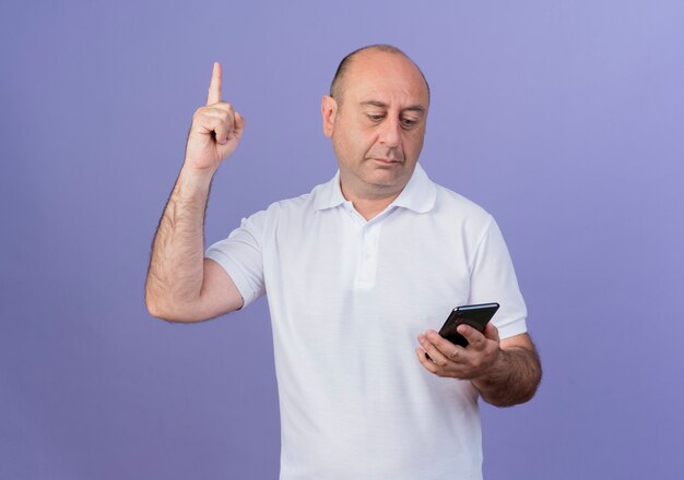 Impresionado empresario maduro casual sosteniendo y mirando el teléfono móvil levantando el dedo aislado sobre fondo púrpura