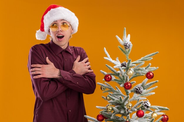 Impresionado y frío joven rubio con gorro de Papá Noel y gafas de pie cerca del árbol de Navidad decorado sobre fondo naranja