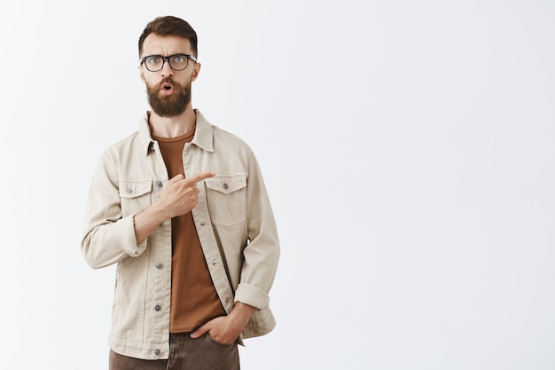 Impresionado guapo barbudo con gafas posando contra la pared blanca