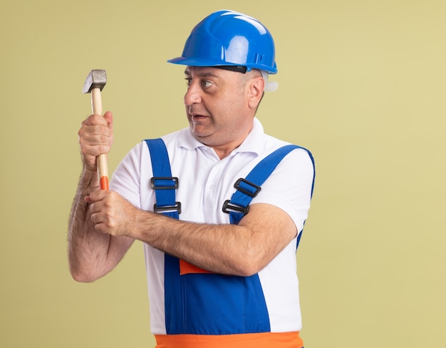 Impresionado hombre adulto constructor en uniforme sostiene y mira martillo aislado en pared verde oliva