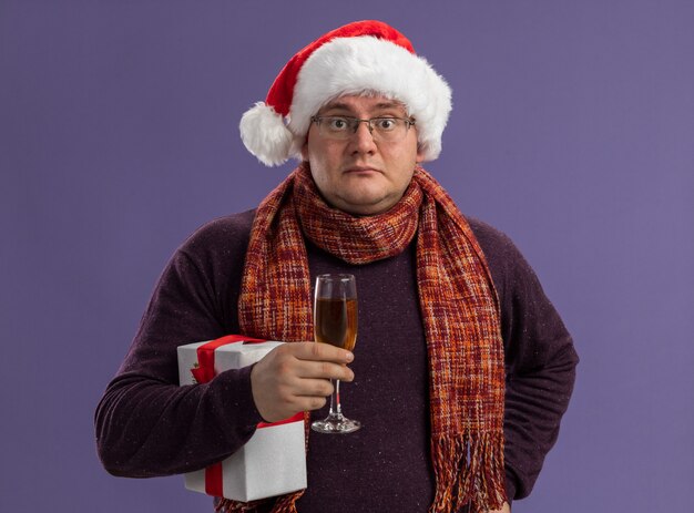 Impresionado hombre adulto con gafas y gorro de Papá Noel con bufanda alrededor del cuello sosteniendo una copa de champán con paquete de regalo manteniendo la mano en la cintura aislada en la pared púrpura