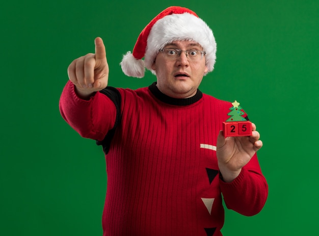 Impresionado hombre adulto con gafas y gorro de Papá Noel con juguete de árbol de Navidad con fecha mirando y apuntando a cámara aislada sobre fondo verde