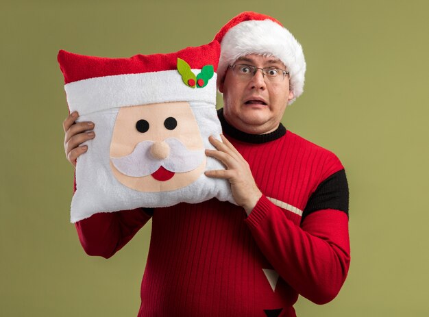 Impresionado hombre adulto con gafas y gorro de Papá Noel sosteniendo la almohada de santa claus por detrás aislado en la pared verde oliva