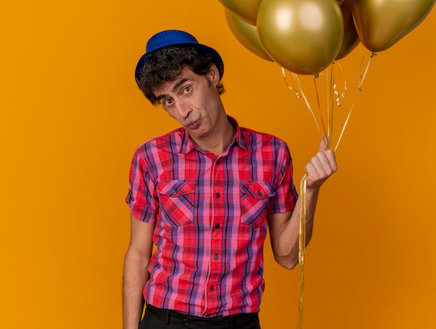 Impresionado hombre caucásico de mediana edad con gorro de fiesta mirando a la cámara sosteniendo globos aislados sobre fondo naranja con espacio de copia