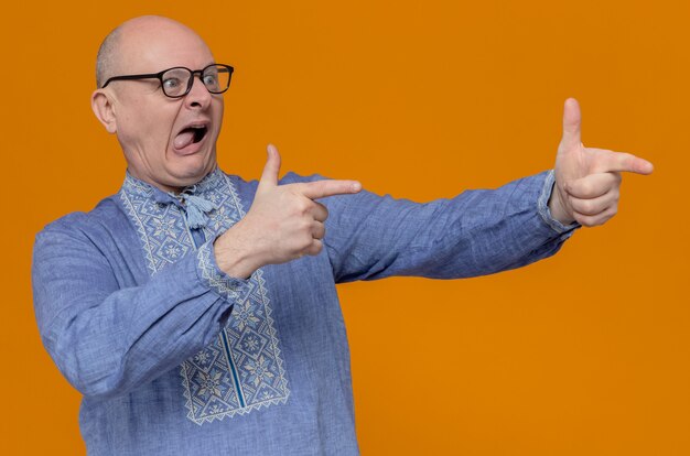 Impresionado hombre eslavo adulto en camisa azul y con gafas ópticas mirando y apuntando a un lado