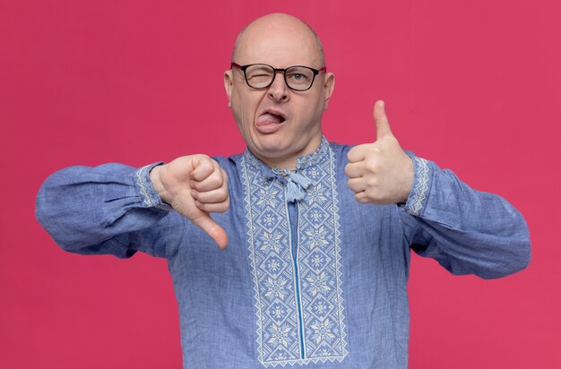 Impresionado hombre eslavo adulto en camisa azul con gafas se pega la lengua con los pulgares hacia arriba y hacia abajo