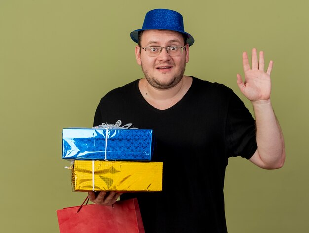 Impresionado hombre eslavo adulto en gafas ópticas con sombrero de fiesta azul se encuentra con la mano levantada sostiene cajas de regalo y bolsa de papel