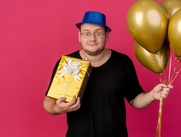 Impresionado hombre eslavo adulto en gafas ópticas con sombrero de fiesta azul sostiene globos de helio y caja de regalo