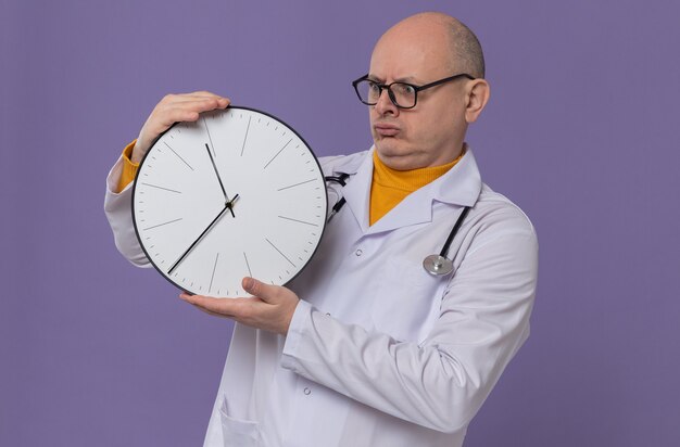 Impresionado hombre eslavo adulto con gafas ópticas en uniforme médico con estetoscopio sosteniendo y mirando el reloj