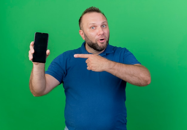 Impresionado hombre eslavo adulto mostrando y apuntando al teléfono móvil aislado en la pared verde