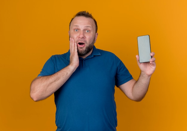 Impresionado hombre eslavo adulto mostrando teléfono móvil manteniendo la mano en la mejilla