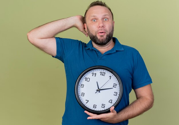 Impresionado hombre eslavo adulto sosteniendo el reloj poniendo la mano detrás de la cabeza