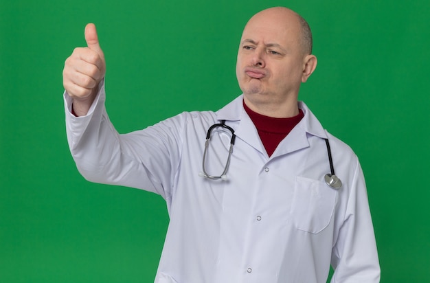 Impresionado hombre eslavo adulto en uniforme médico con estetoscopio pulgar hacia arriba mirando de lado