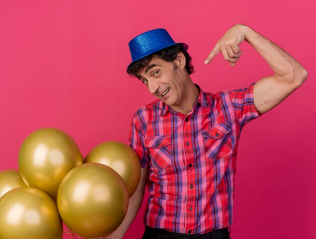 Impresionado hombre de fiesta de mediana edad con sombrero de fiesta mirando al frente sosteniendo y apuntando a globos aislados en la pared carmesí