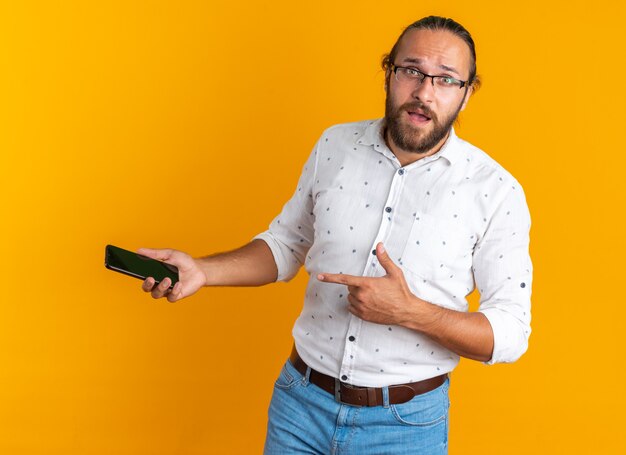 Impresionado hombre guapo adulto con gafas mirando a la cámara sosteniendo y apuntando al teléfono móvil aislado en la pared naranja