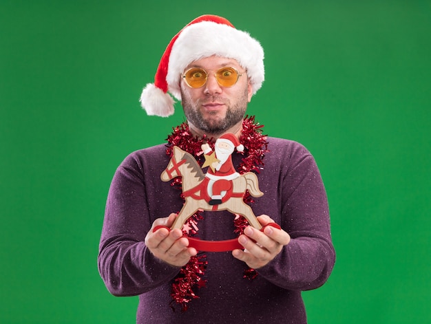 Impresionado hombre de mediana edad con gorro de Papá Noel y guirnalda de oropel alrededor del cuello con gafas estirando santa en estatuilla de caballo mecedora aislada en la pared verde