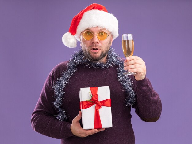 Impresionado hombre de mediana edad con gorro de Papá Noel y guirnalda de oropel alrededor del cuello con gafas con paquete de regalo y copa de champán aislado en la pared púrpura