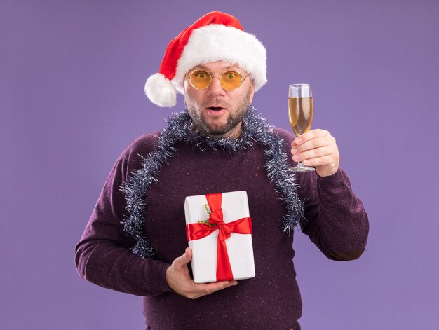 Impresionado hombre de mediana edad con gorro de Papá Noel y guirnalda de oropel alrededor del cuello con gafas con paquete de regalo y copa de champán aislado en la pared púrpura