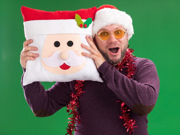 Foto gratuita impresionado hombre de mediana edad con gorro de papá noel y guirnalda de oropel alrededor del cuello con gafas sosteniendo la almohada de santa claus tocando la cabeza con ella aislada en la pared verde