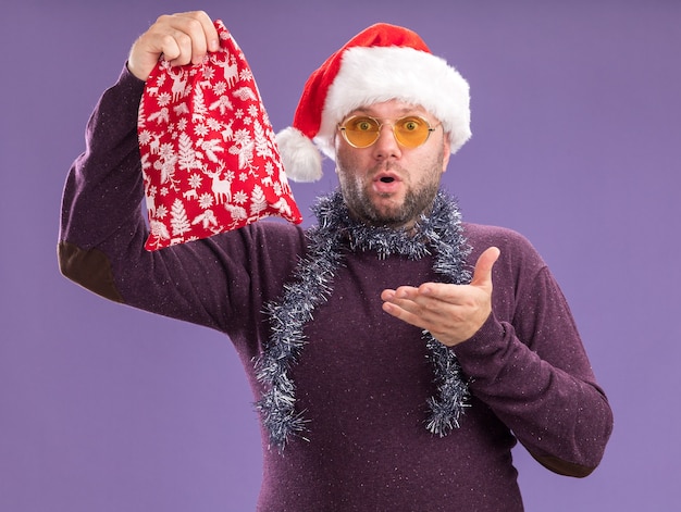 Impresionado hombre de mediana edad con gorro de Papá Noel y guirnalda de oropel alrededor del cuello con gafas sosteniendo y apuntando al saco de regalo de Navidad mirando a cámara aislada sobre fondo púrpura