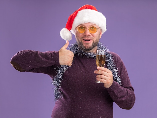 Impresionado hombre de mediana edad con gorro de Papá Noel y guirnalda de oropel alrededor del cuello con gafas sosteniendo una copa de champán mostrando el pulgar hacia arriba aislado en la pared púrpura