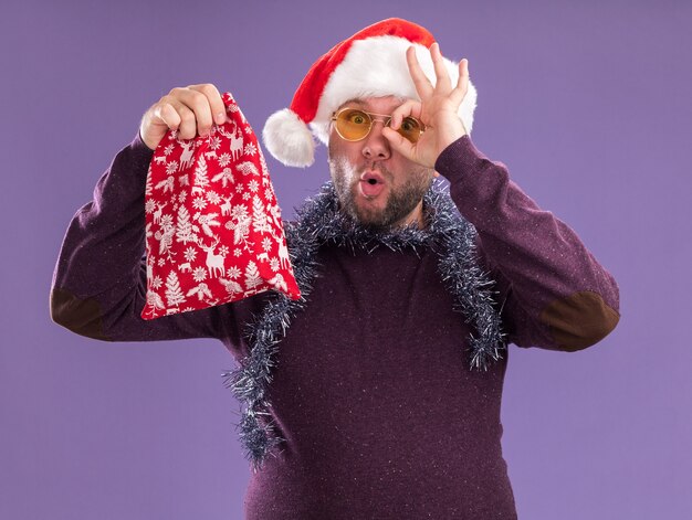 Impresionado hombre de mediana edad con gorro de Papá Noel y guirnalda de oropel alrededor del cuello con gafas sosteniendo un saco de regalo de Navidad mirando a la cámara haciendo gesto de mirada aislado sobre fondo púrpura