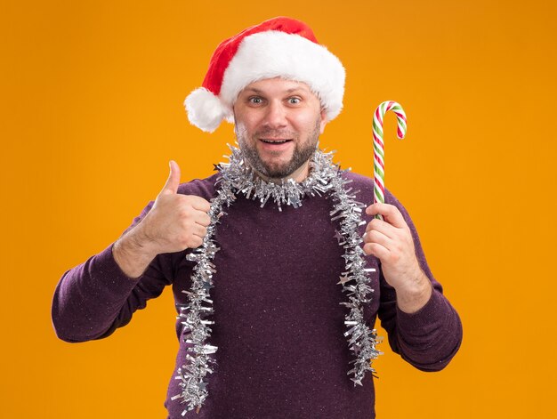 Impresionado hombre de mediana edad con gorro de Papá Noel y guirnalda de oropel alrededor del cuello sosteniendo bastón dulce de Navidad mirando a cámara mostrando el pulgar hacia arriba aislado sobre fondo naranja