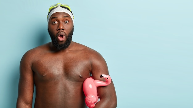 Impresionado hombre negro con cuerpo fuerte y musculoso, pasa el tiempo libre haciendo su pasatiempo favorito, usa gafas y gorro de baño, sostiene un anillo de goma en forma de flamenco, sorprendido por el frío estropeado