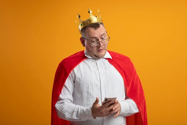 Impresionado hombre superhéroe adulto en capa roja con gafas y corona sosteniendo y mirando el teléfono móvil aislado en la pared naranja